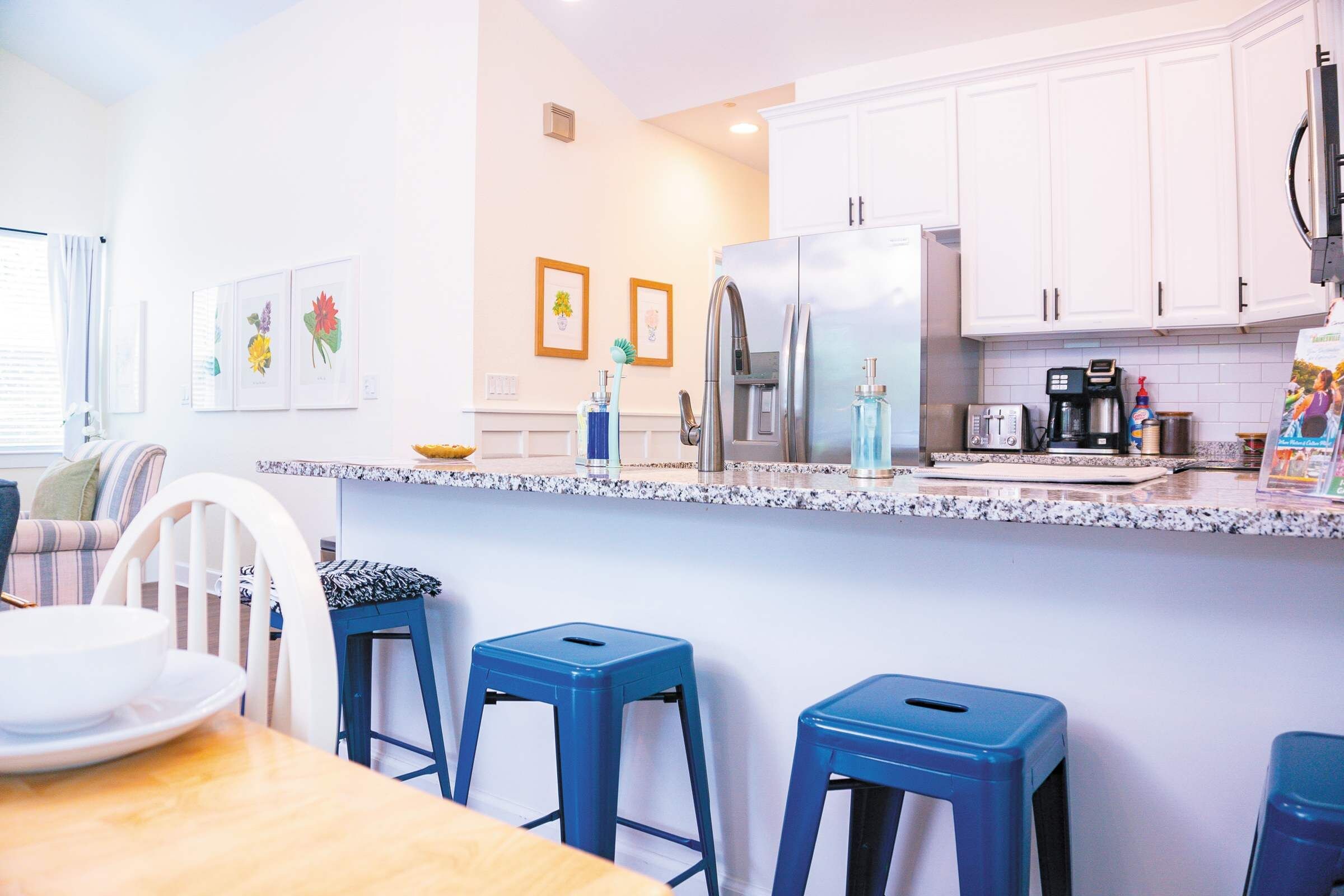 Una cocina moderna con armarios blancos, un refrigerador de acero inoxidable y una encimera de granito. Hay tres taburetes azules y una mesa de comedor con un plato blanco.