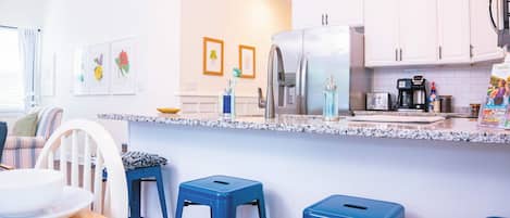 Una cocina moderna con armarios blancos, un refrigerador de acero inoxidable y una encimera de granito. Hay tres taburetes azules y una mesa de comedor con un plato blanco.