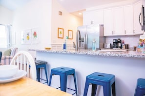 Cozinha moderna com armários brancos, geladeira de aço inoxidável e bancada de granito. Há três banquetas azuis e uma mesa de jantar com um prato branco.