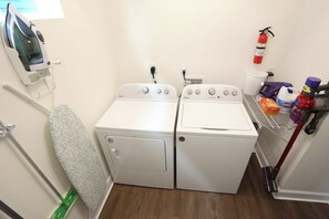 A laundry room with two front-loading washing machines, a wall-mounted iron, a mop, a fire extinguisher, and cleaning supplies. - The Prairie Suite. Newly Built, Upscale, Central (Gainesville)