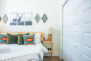A neatly made bed with patterned pillows, a bedside table with a lamp, and wall art including a deer and geometric designs. - The Prairie Suite. Newly Built, Upscale, Central (Gainesville)