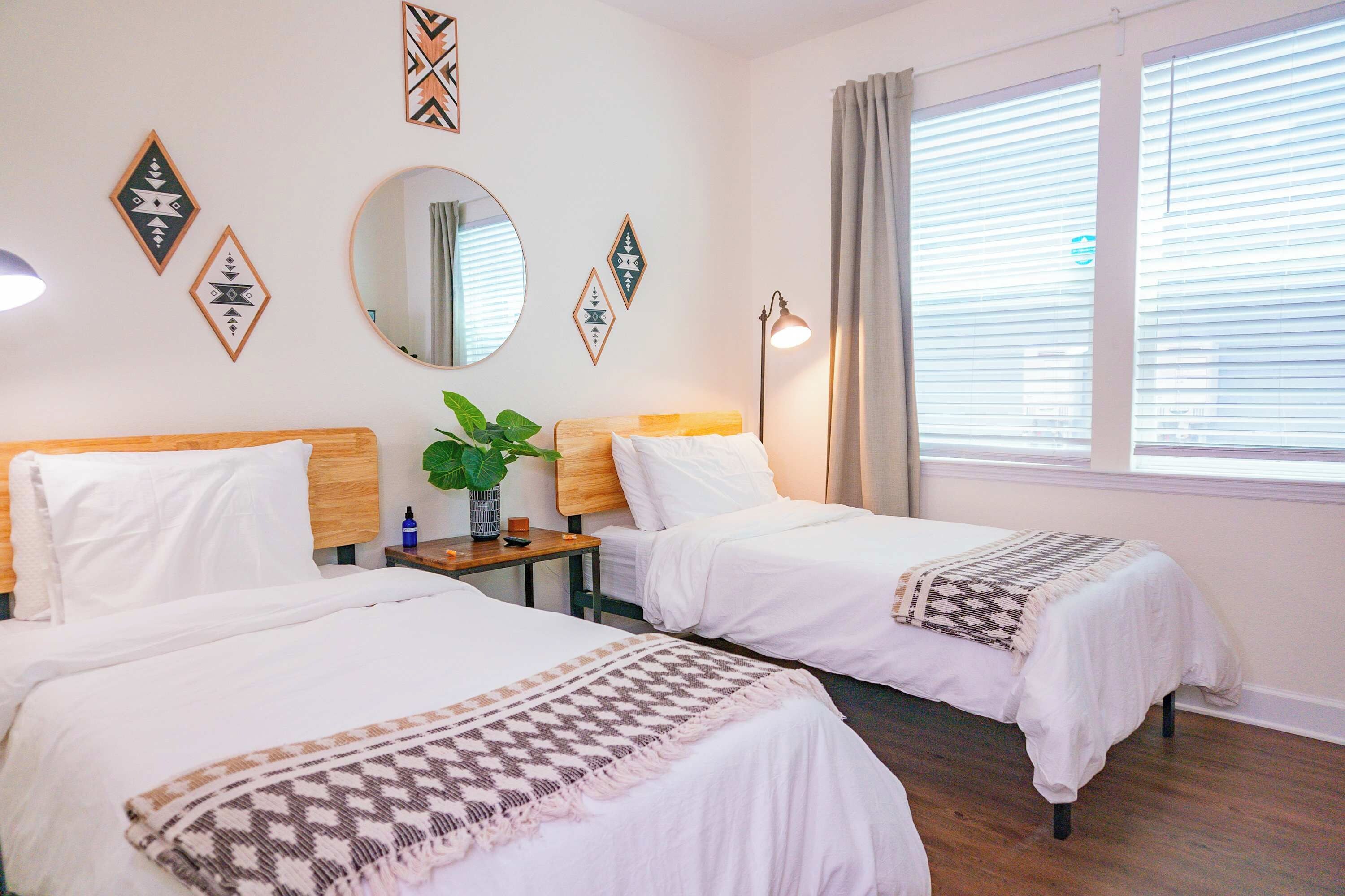 A hotel room with two beds, a round mirror, decorative wall art, a plant, and a window with curtains.