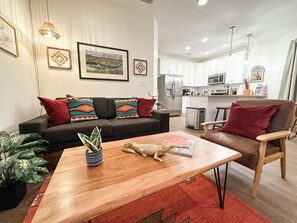 A modern living room with a sofa, coffee table, and decorative pillows. A kitchen with white cabinets and appliances is visible in the background. - The Prairie Suite. Newly Built, Upscale, Central (Gainesville)