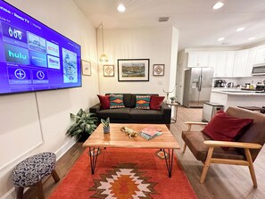A modern living room with a flat-screen TV displaying streaming services, a black sofa with colorful cushions, a wooden coffee table, and a small wooden chair. - The Prairie Suite. Newly Built, Upscale, Central (Gainesville)