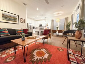 Un salon moderne avec un canapé, une table basse et des fauteuils, un coin repas avec une table et des chaises, et une cuisine en arrière-plan.