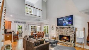 A spacious living room with a stone fireplace, a television mounted above it, and a dining area with a view of the outdoors.