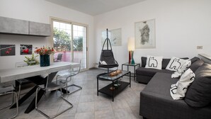A modern living room with a sofa, coffee table, and chairs. There is a window with a view of trees and a balcony. - Casa Marisa by Wonderful Italy (Golfo Aranci)