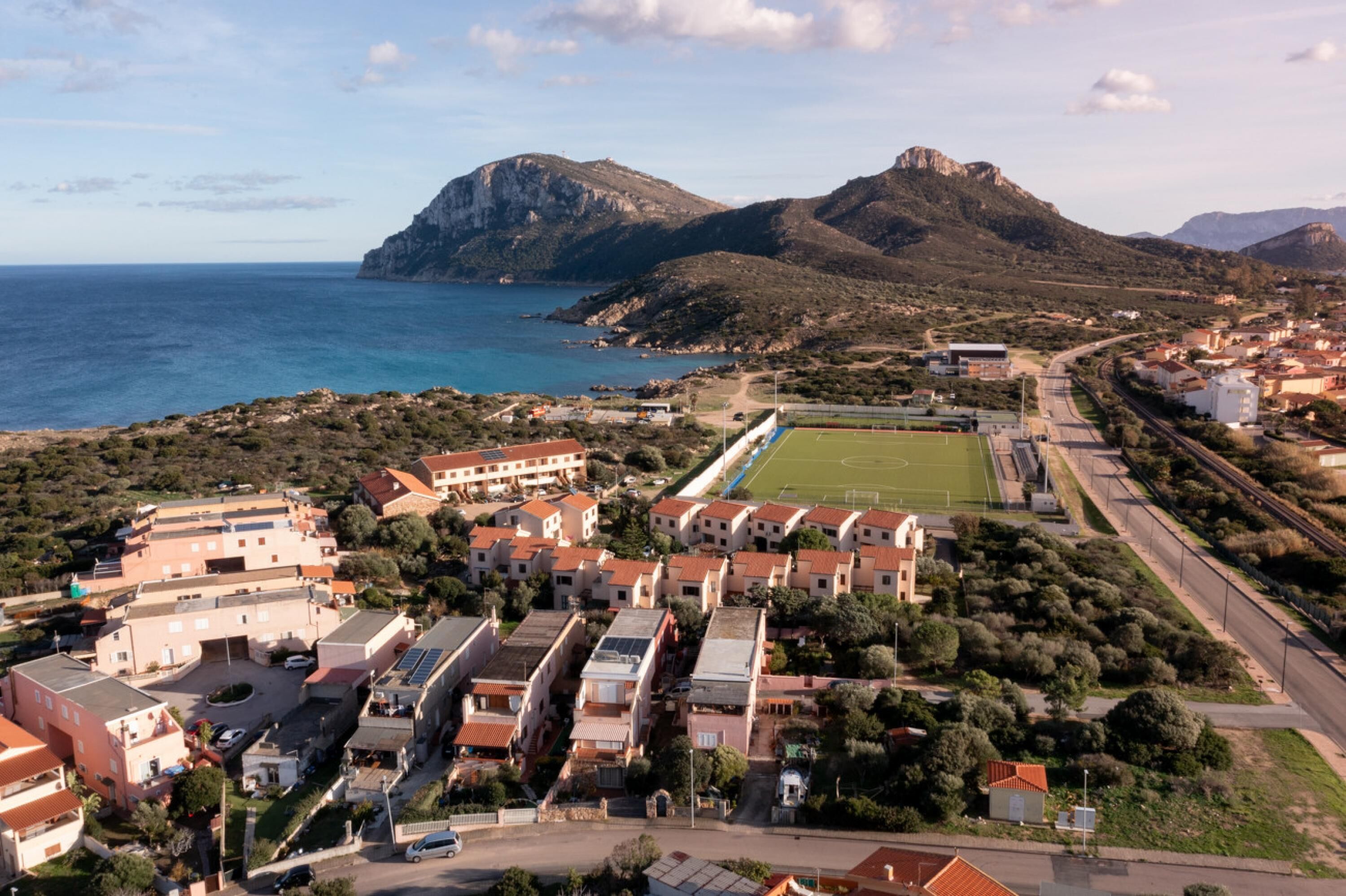 An aerial view of a coastal town with a prominent mountain, a sports field, and residential buildings.