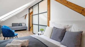 A bedroom with a sloped ceiling, a bed with grey bedding, a blue chair, and a small table with a plant.