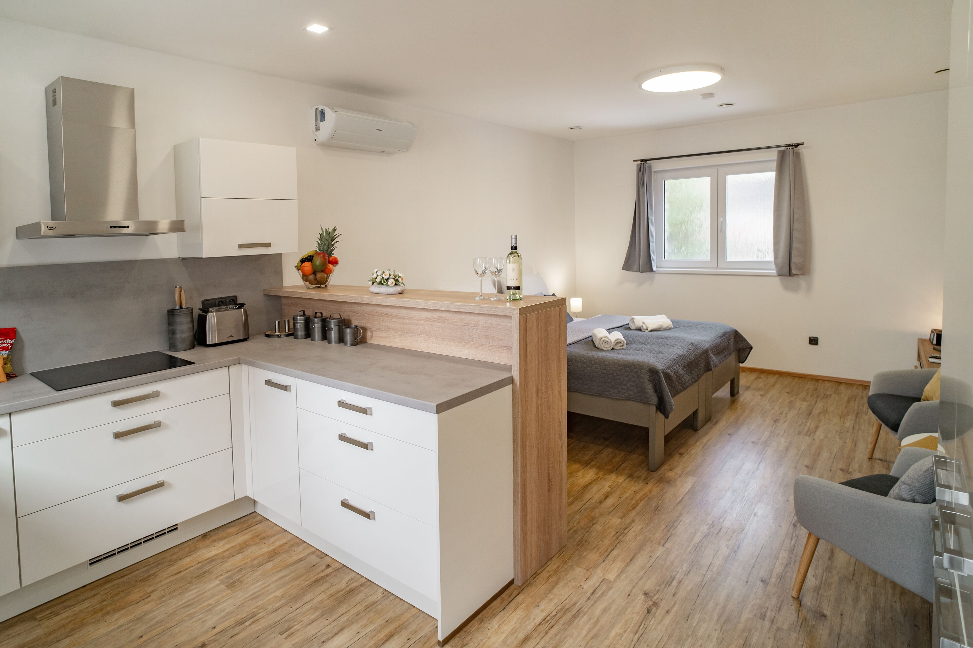 A modern kitchen with white cabinets, a built-in stove, and a wooden countertop. There is a bed with a gray blanket and a window with curtains.