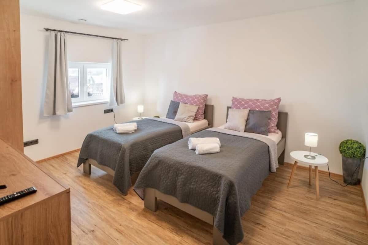 Quarto de hotel com duas camas de solteiro, uma mesinha com candeeiro, janela com cortinas e chão de madeira.