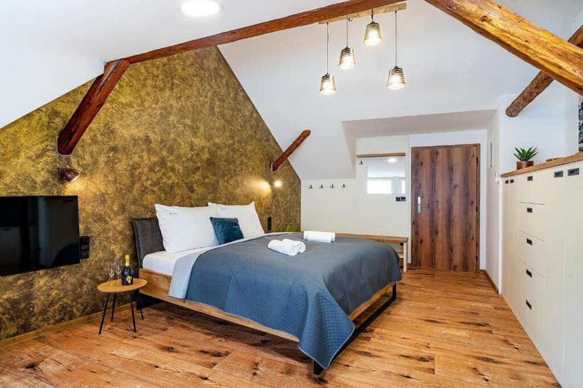 Um quarto moderno com teto inclinado, vigas de madeira, uma cama com colcha azul, uma mesinha com uma garrafa e copos, e uma televisão fixada na parede.