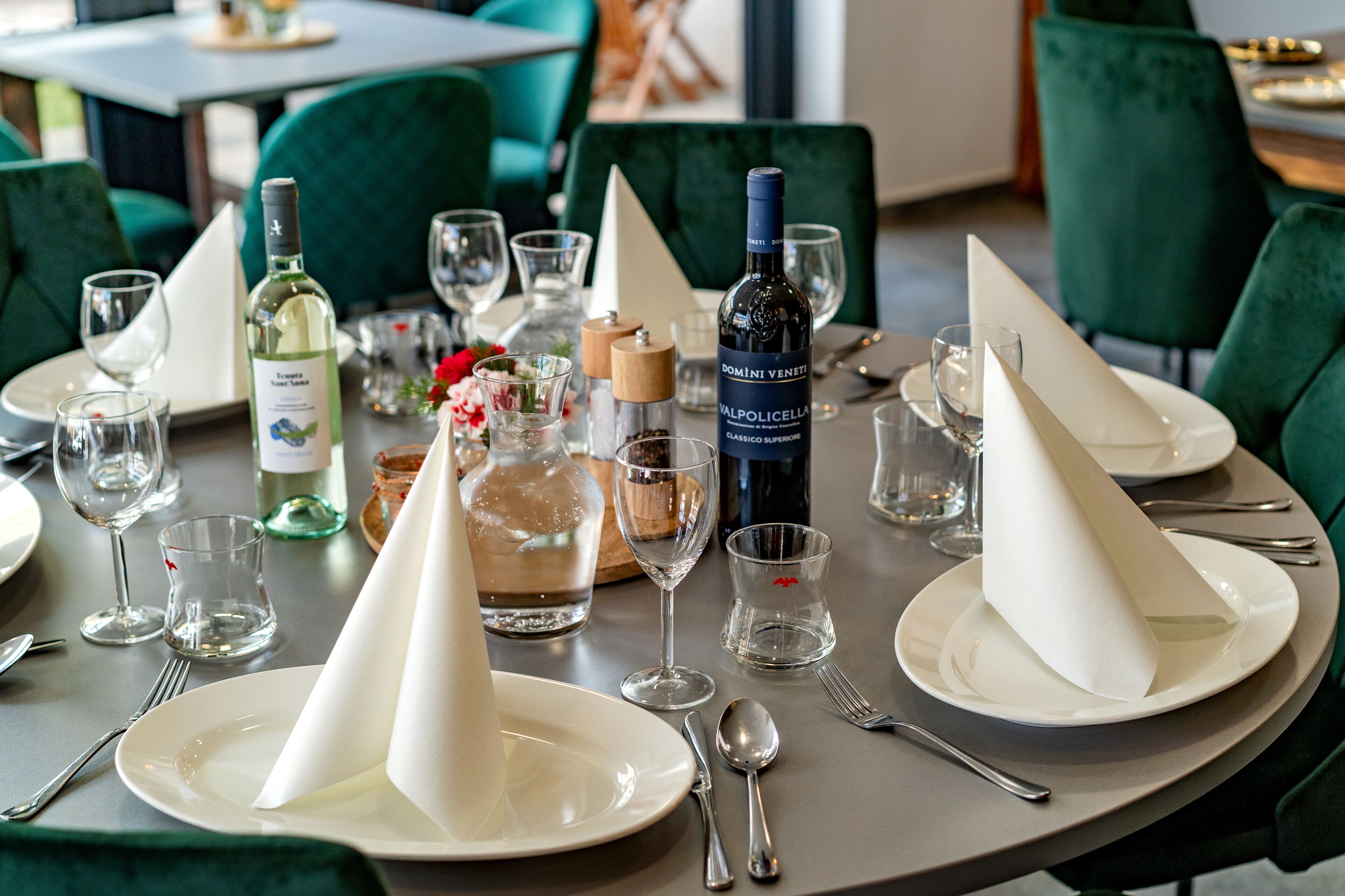A dining table set with white plates, silverware, and wine glasses, surrounded by green upholstered chairs.