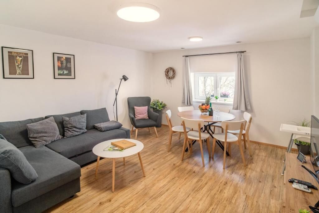 Una sala de estar moderna con un sofá gris, una mesa de comedor redonda con sillas, una mesita blanca y un televisor sobre un mueble.