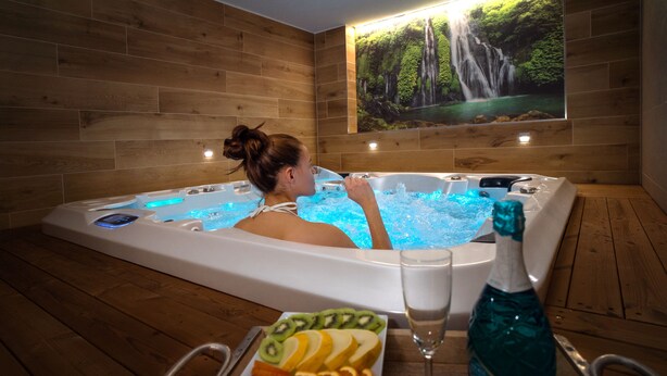 A person relaxing in a hot tub with a waterfall mural on the wall, surrounded by fruit and a bottle of blue liquid.