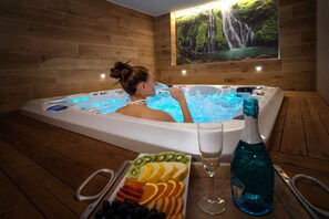 A person relaxing in a hot tub with a waterfall mural on the wall, surrounded by fruit and a bottle of blue liquid. - HOTEL SMRŽ (Sepekov)