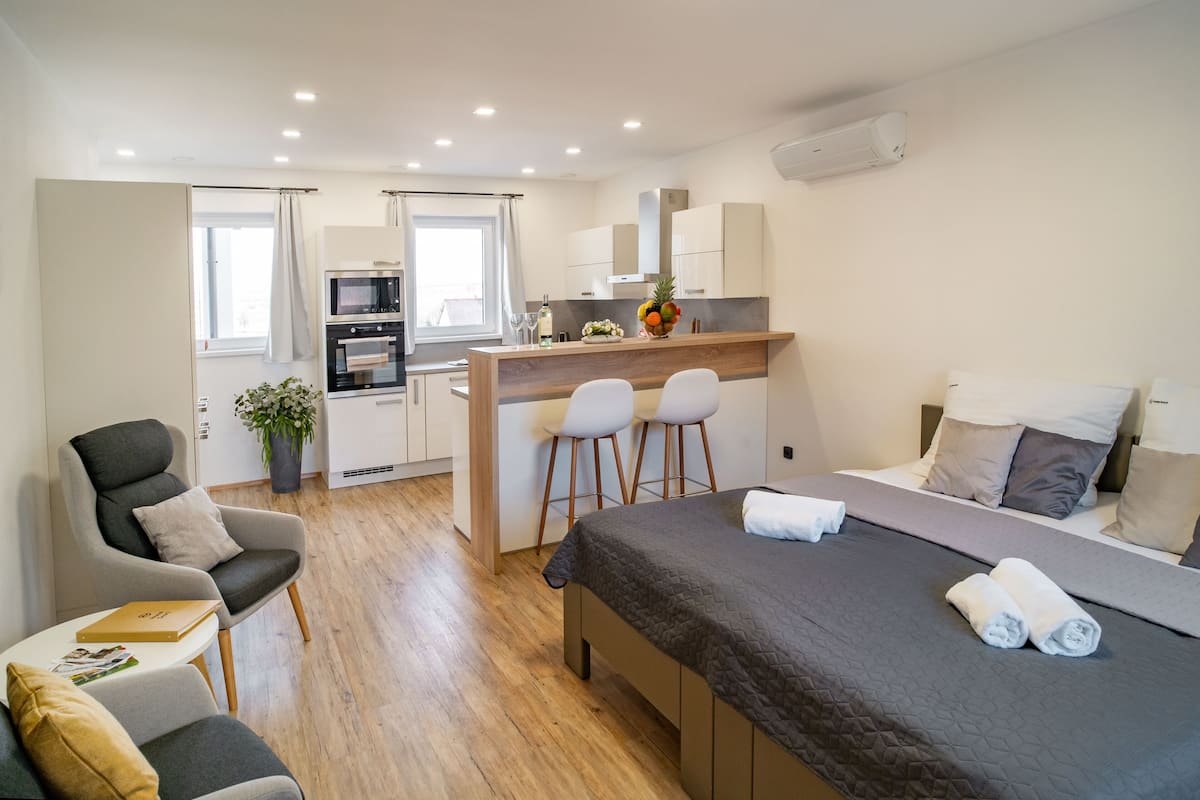 Quarto de hotel moderno com cama, escrivaninha e duas cadeiras, cozinha compacta e uma janela grande.