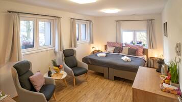 Et moderne hotelværelse med en seng, to lænestole, et lille bord med en flaske og frugt, samt et skrivebord med en lampe og planter.