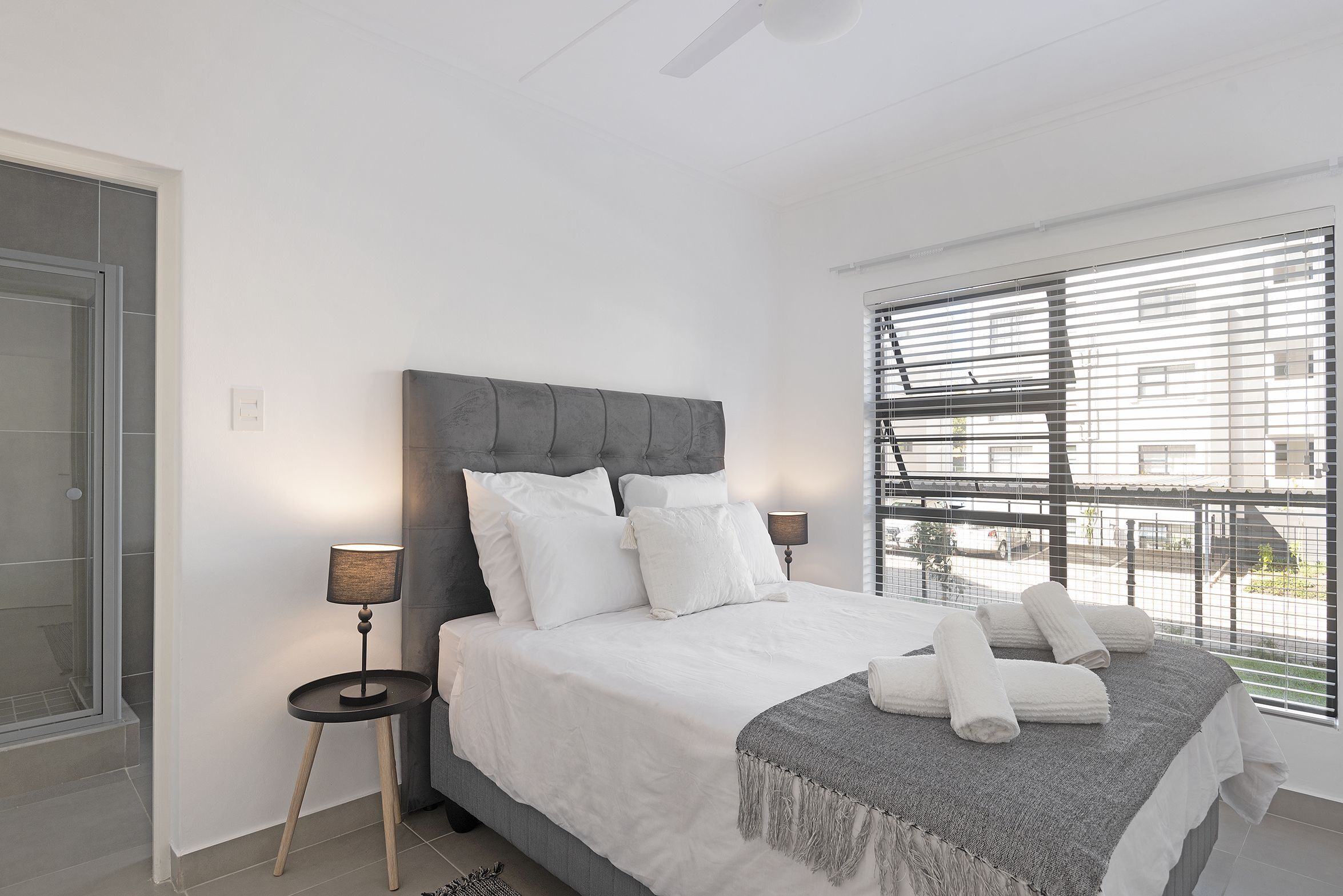 A modern bedroom with a large bed, two bedside tables, a grey headboard, and a view of the street through large windows.