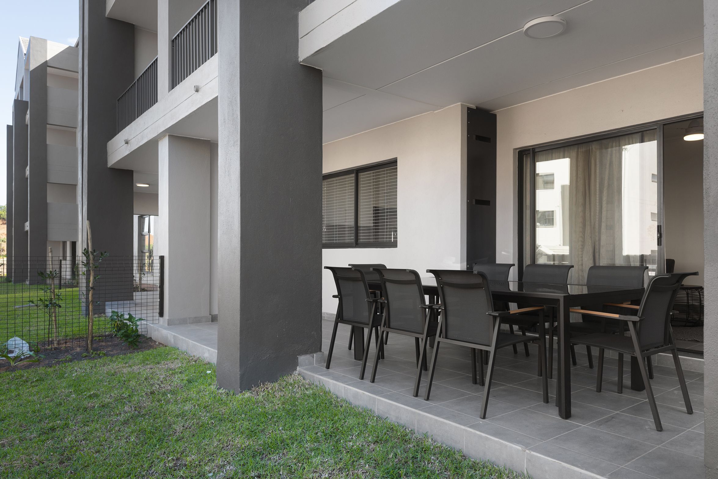 A modern outdoor patio with a dining table and chairs, surrounded by a low fence and greenery.