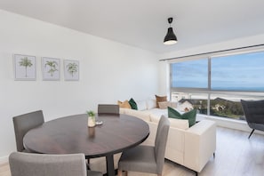 A modern living room with a round dining table, grey upholstered chairs, a white sofa with green and brown cushions, and a large window offering a view of the city. - Blouberg Heights 207 (Cape Town)