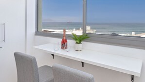 Une chambre moderne avec une grande fenêtre donnant sur l’océan, un banc blanc, deux fauteuils gris, une petite plante en pot et une bouteille de vin.