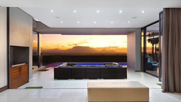 A modern living room with a large window overlooking a sunset, a sofa, and a small pool area. - Baronetcy Kings Villa (Cape Town)