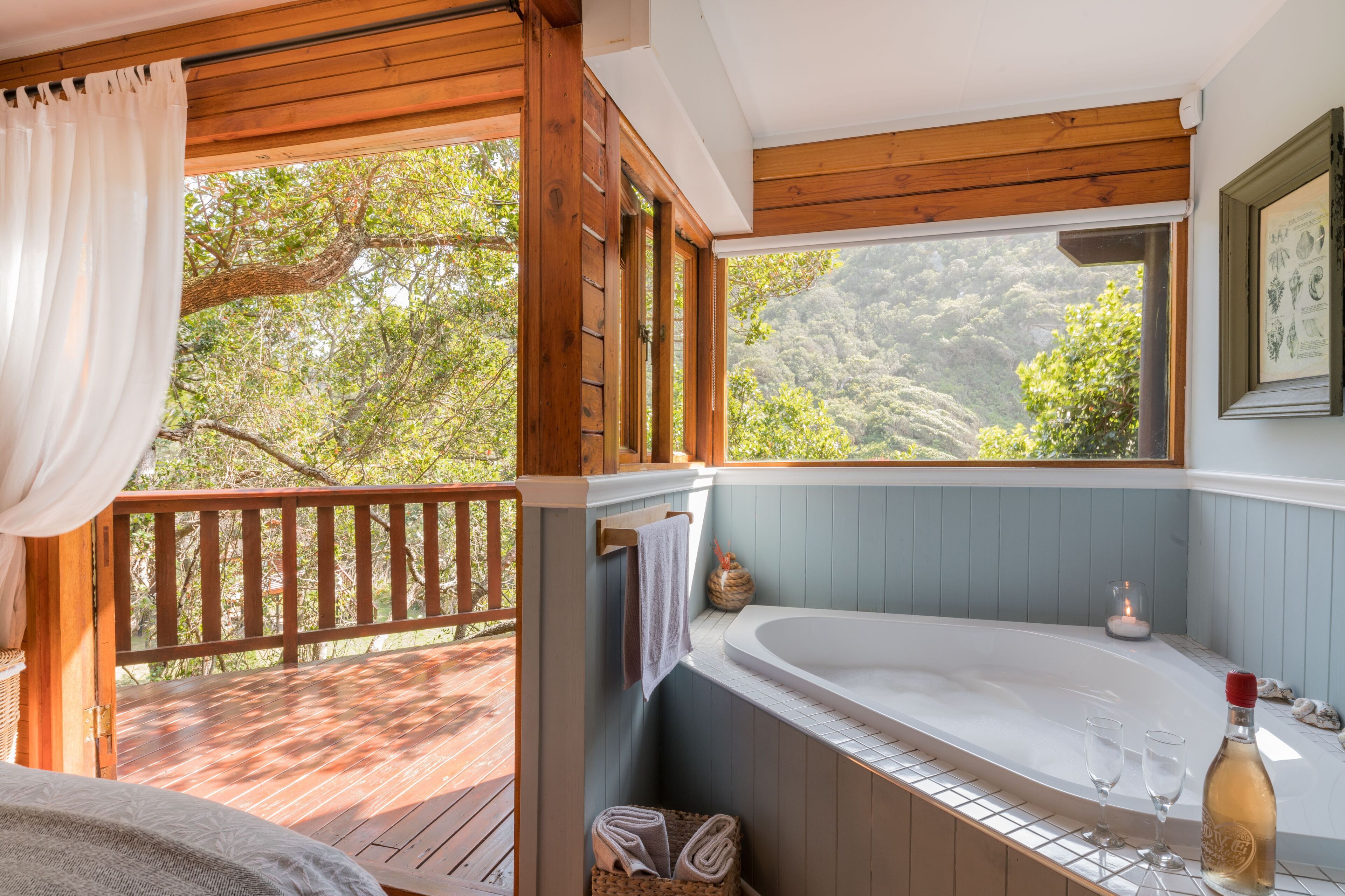 A wooden deck with a view of trees, a wooden cabin, a large bathtub, and a bottle of wine.