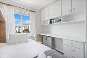 Un dormitorio con cama, cortinas blancas, una ventana con vistas, un escritorio con libros y un radiador.