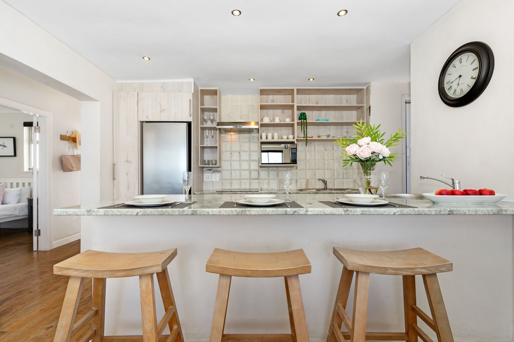 A modern kitchen with a central island, bar stools, a clock, and a dining area with a sofa.