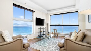 A modern living room with a glass coffee table, beige sofas, a TV mounted on the wall, and large windows offering a view of the ocean.