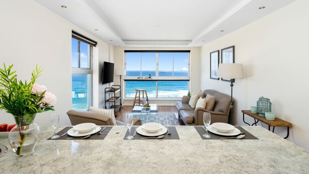 A modern living room with a large window overlooking the ocean, a sofa, a coffee table, and a dining area set for two.