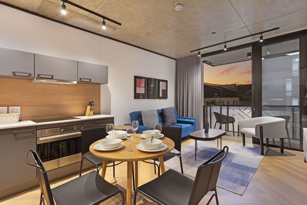 A modern kitchen with stainless steel appliances, a dining area with a round table and chairs, a blue sofa, and a balcony with a view of the sunset.