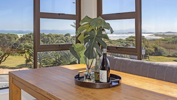 Una mesa de madera con una botella de vino y una planta sobre una bandeja, con vistas a un paisaje pintoresco a través de amplios ventanales.