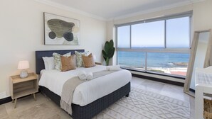 A bedroom with a large bed, a nightstand, a potted plant, and a view of the ocean. - Blouberg Heights 603 (Cape Town)