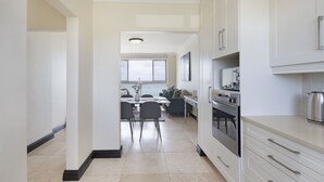 A modern kitchen with white cabinets, a stainless steel oven, and a dining area with a table and chairs. - Blouberg Heights 603 (Cape Town)