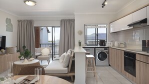 A modern apartment with a kitchenette, a dining area, a living room with a sofa, and a balcony with curtains.