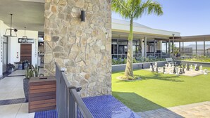 A stone facade with a restaurant sign, a palm tree, and a chessboard in a courtyard.