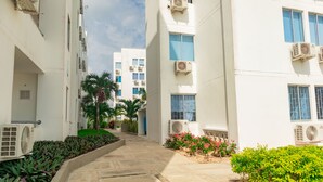 Un edificio residencial con varias unidades de aire acondicionado, un sendero pavimentado y algo de vegetación.