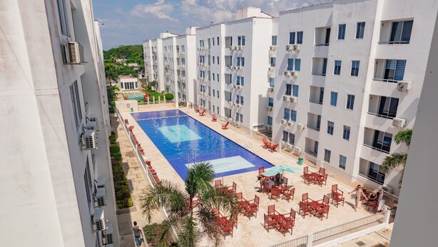 Zona de piscina en la azotea con varios niveles de edificios blancos, palmeras y sillones reclinables rojos.