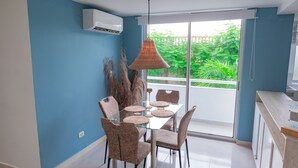 Un comedor con una mesa y sillas de vidrio, aire acondicionado y vista a zonas verdes a través de una puerta corredera de vidrio.