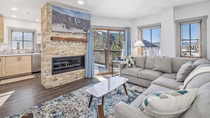 A modern living room with a fireplace, a sofa, a coffee table, and a view of a snowy mountain landscape through large windows.