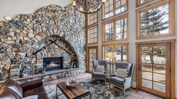 A stone fireplace, a leather sofa, and a chandelier in a room with large windows.