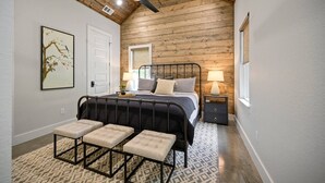 Un dormitorio con techo de madera, una cama con cabecera, dos mesitas de noche con lámparas y un banco con cojines.