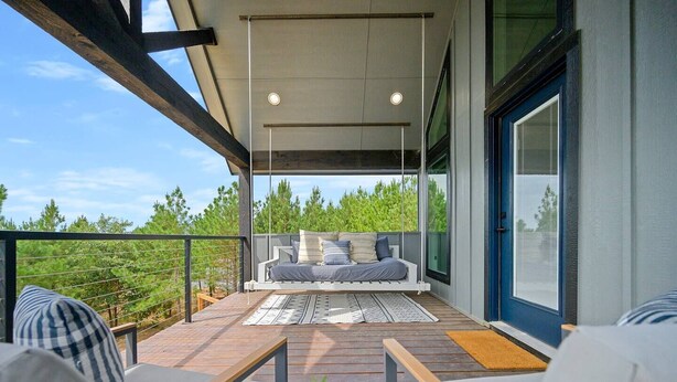 Un área de estar exterior moderna con una cama suspendida, piso de madera y vista a árboles.