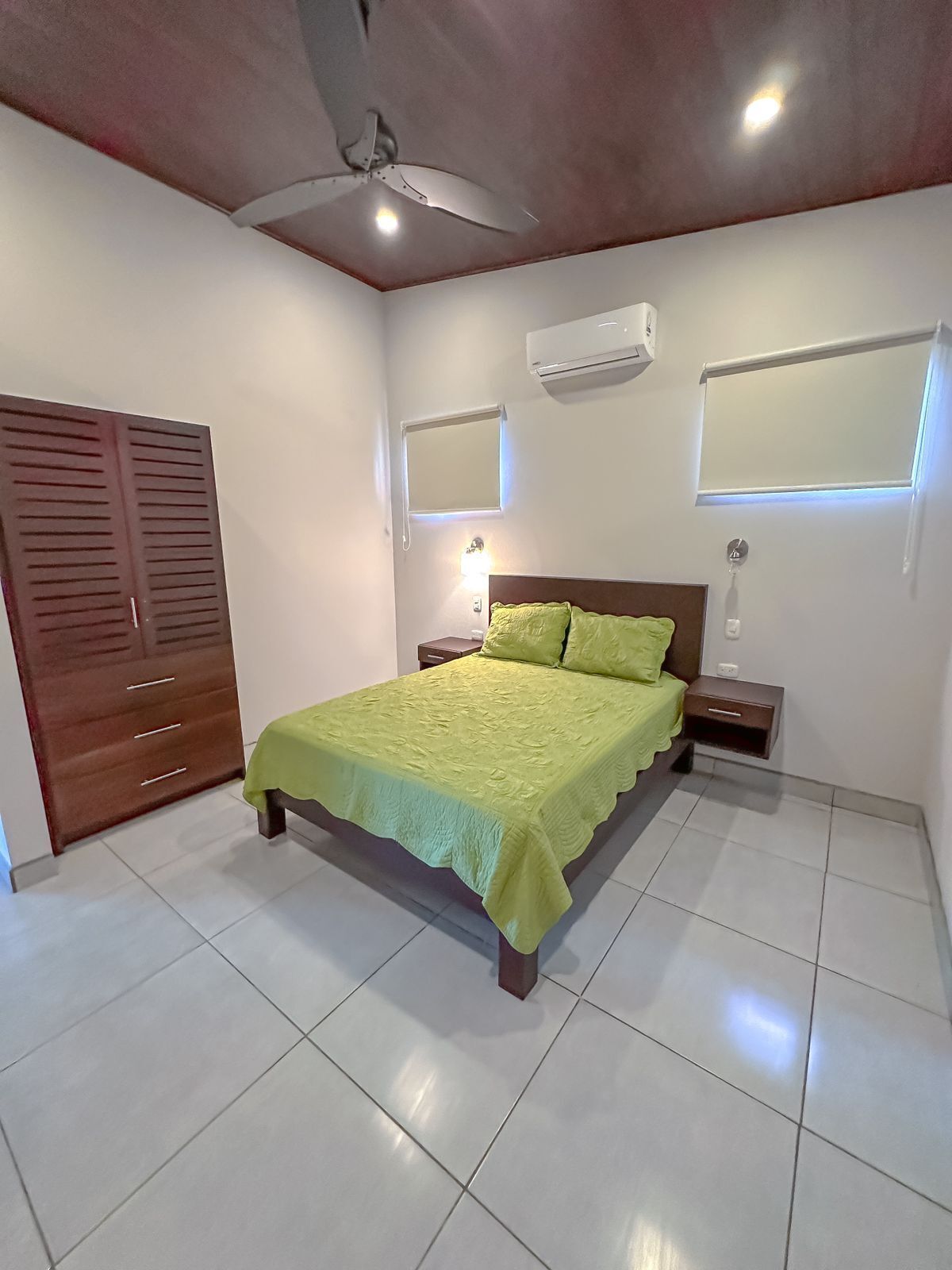 Un dormitorio con cama, ventilador de techo, equipo de aire acondicionado, armario y dos mesitas de noche con lámparas.