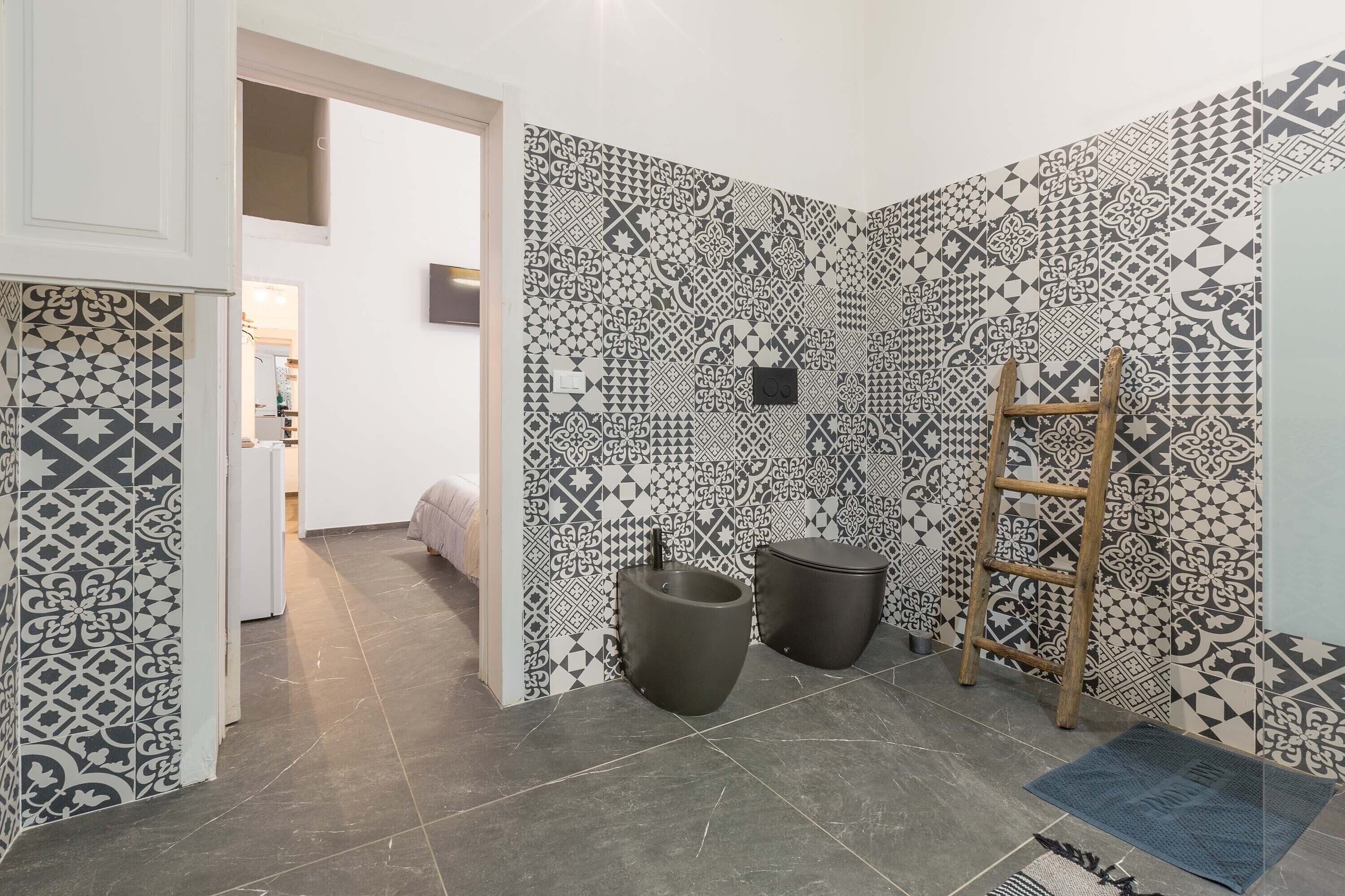 Ein Badezimmer mit gemusterten Fliesen, zwei schwarzen Waschbecken, einer Holzleiter und einer blauen Matte.