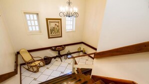 A wooden staircase with a handrail, a wicker chair, a small table with a vase, and two windows with white frames. - Pousada Casa Gialla - Campos do Jordão (Campos do Jordão)