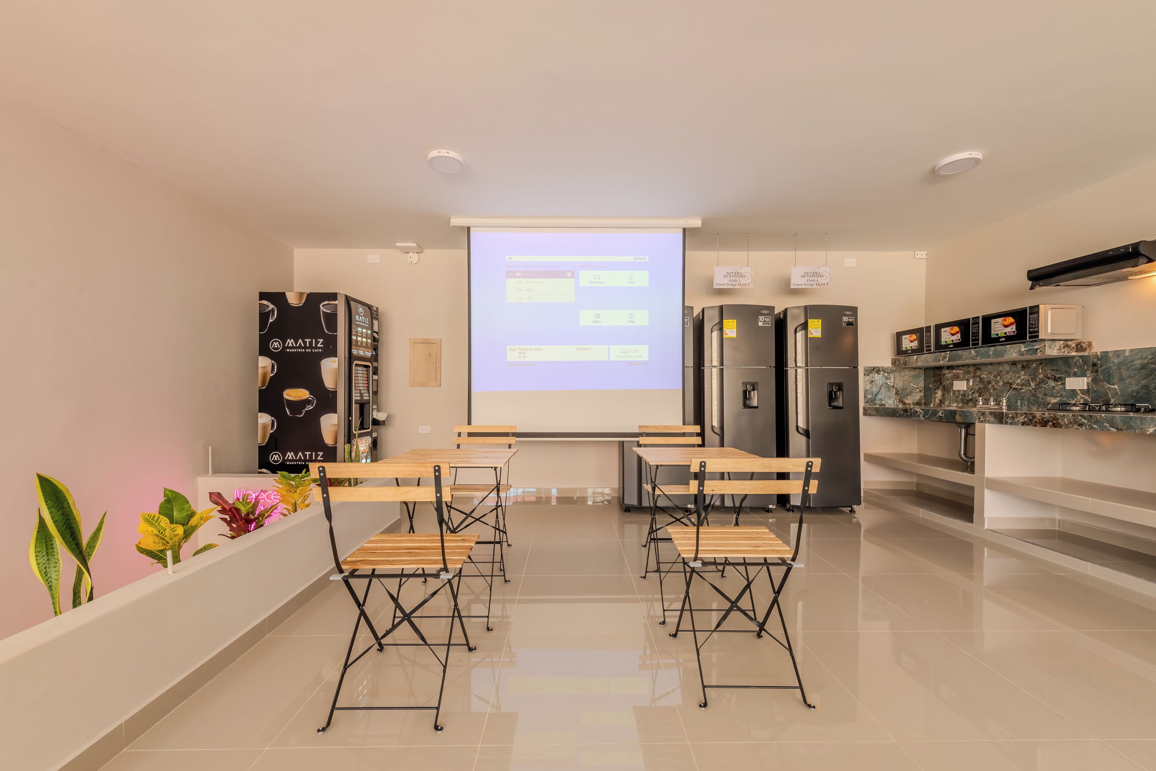 Una sala de conferencias moderna con pantalla de proyector, mesas, sillas, una kitchenette y una pared vegetal.