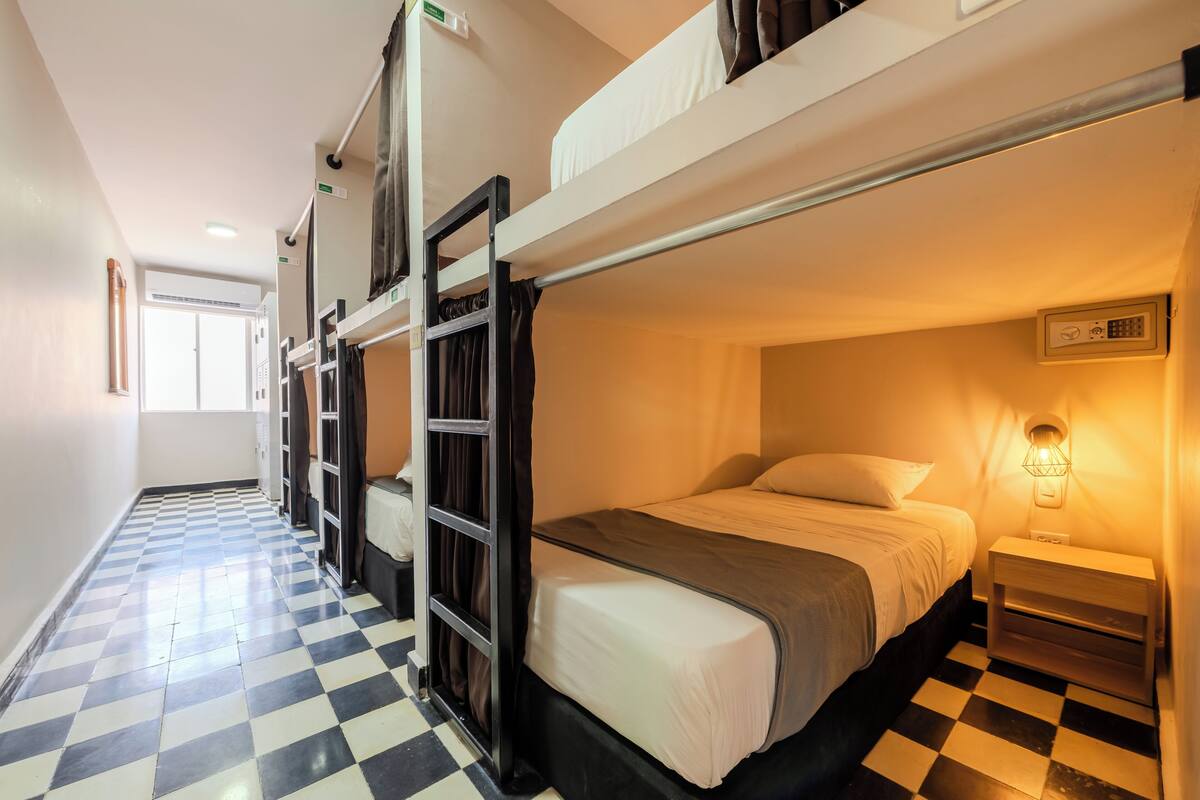 A bunk bed room with a checkered floor, a bedside table with a lamp, and a door leading to another room.